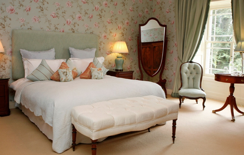 A Tankardstown bedroom with floral wallpaper, a dressed bed, lamps, and an upholstered chair by the window.