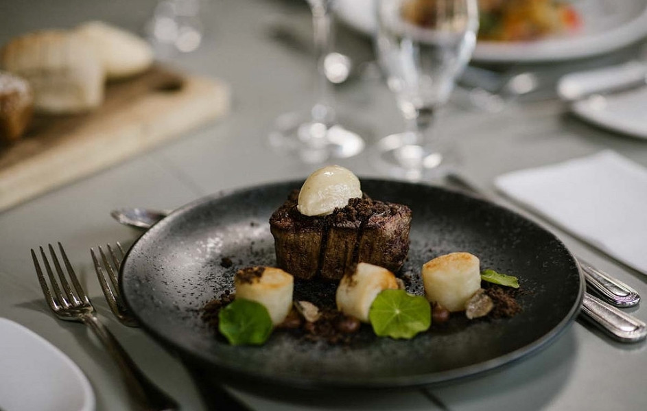 A plated dish on a dark plate with glasses and table settings nearby.