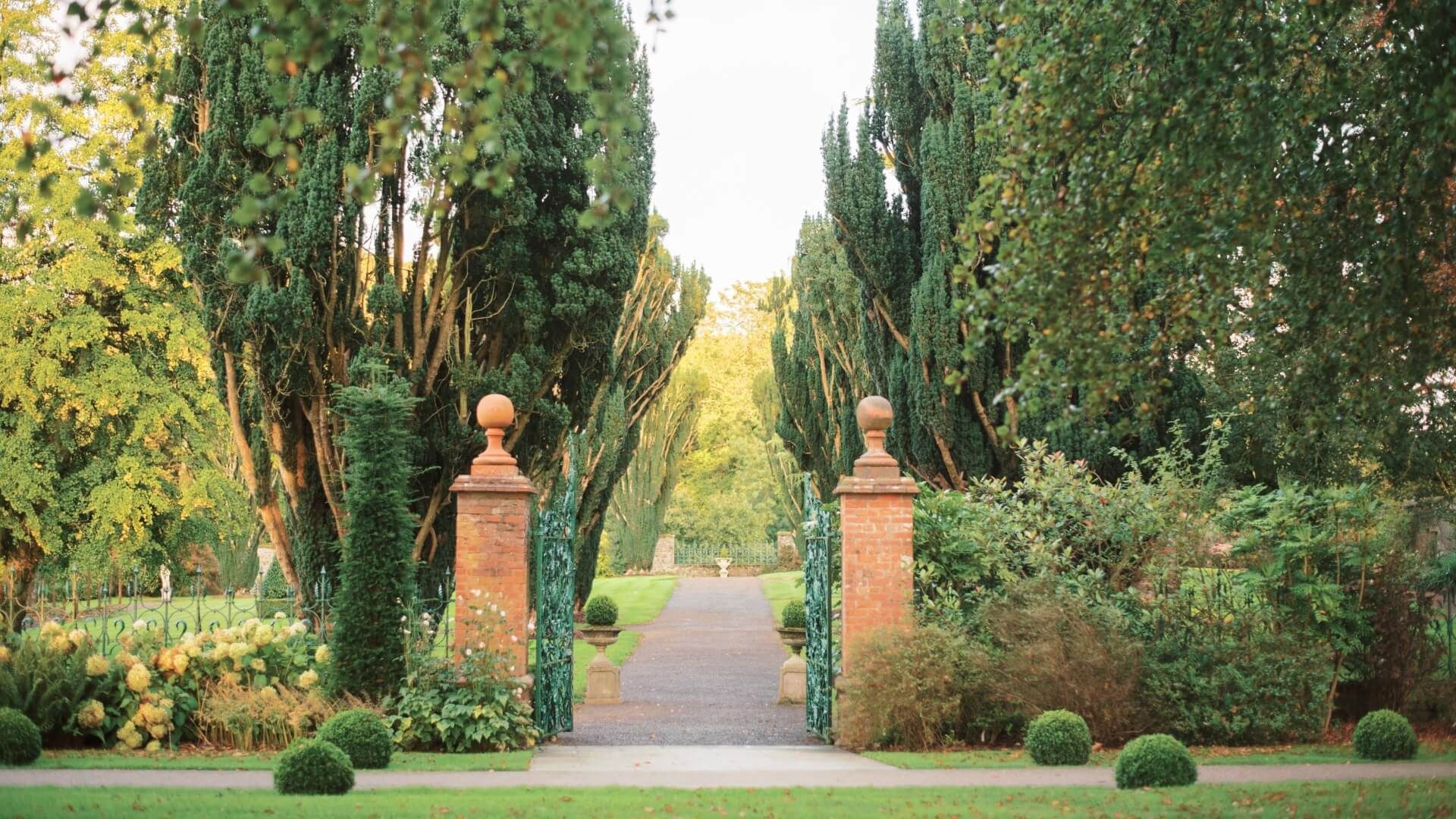 A formal garden entrance with brick gateposts, iron gates, clipped planting, and tall trees.